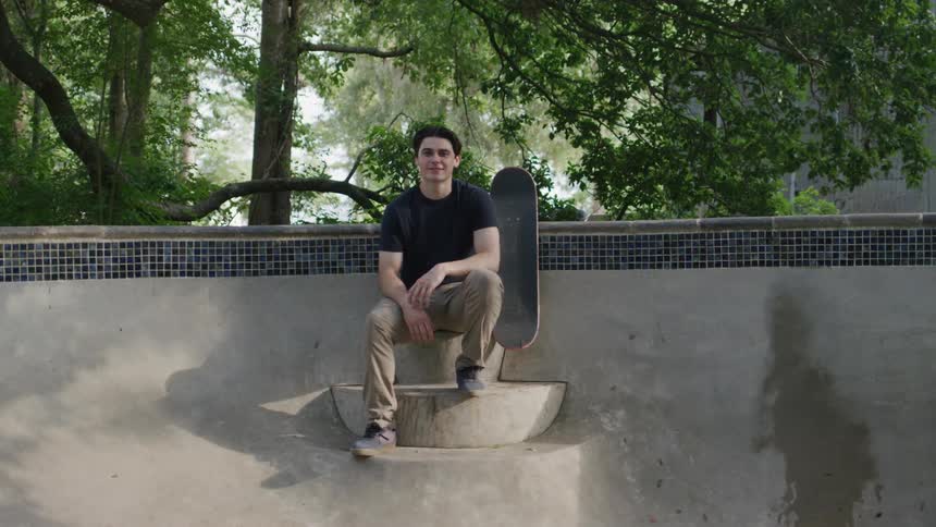A young man sits in a public park with a skateboard beside him at midday