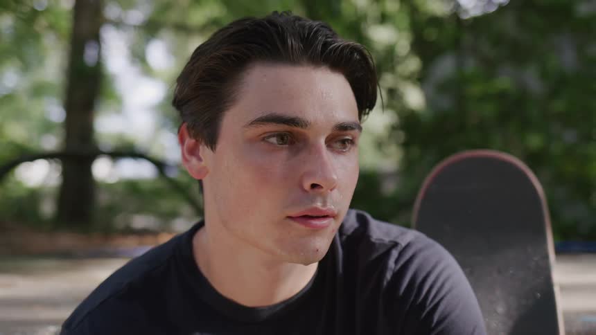 A young man sits contemplatively in an outdoor park or backyard setting with a skateboard resting beside him as he looks down and then up with a serious expression