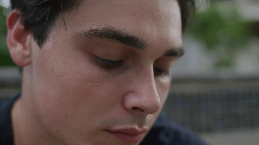 The video shows a closeup of a young man with dark hair looking down with a serious expression the background is blurred focusing attention on his face