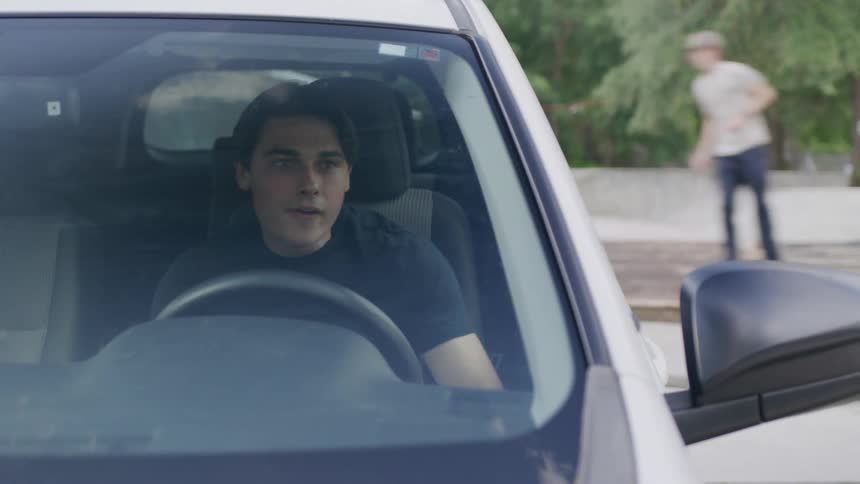 A teenager sits inside his car operating his phone in a public park while other kids ride their skateboards at midday
