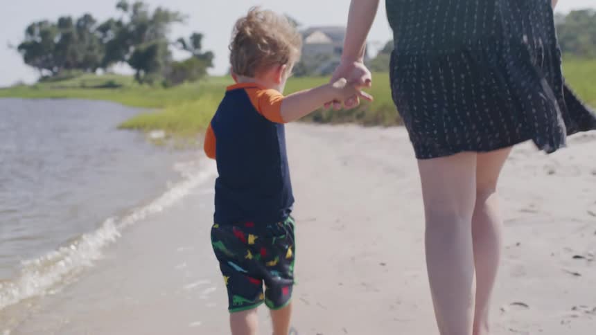 A woman holds a young boys hand as they walk along the shoreline on a sunny day