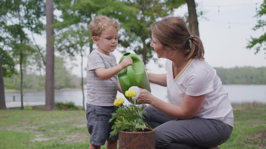 A woman and a small boy crouch next to a potted plant and the boy pours water from a green watering can onto a small yellow flower
