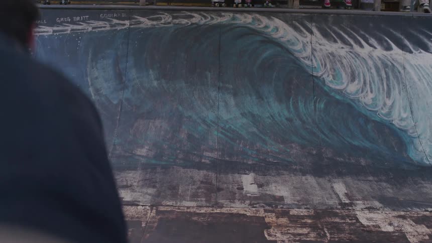 A man skateboards back and forth on a half pipe ramp that is painted with a large mural of a crashing wave