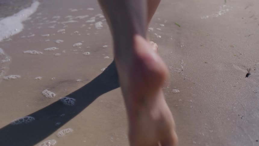A persons bare feet walk along the wet sand at the waters edge