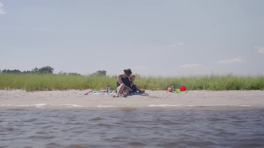 A young mother is sitting on a blanket on a sandy beach next to tall grass she is holding her small child in her lap and there are sand toys nearby