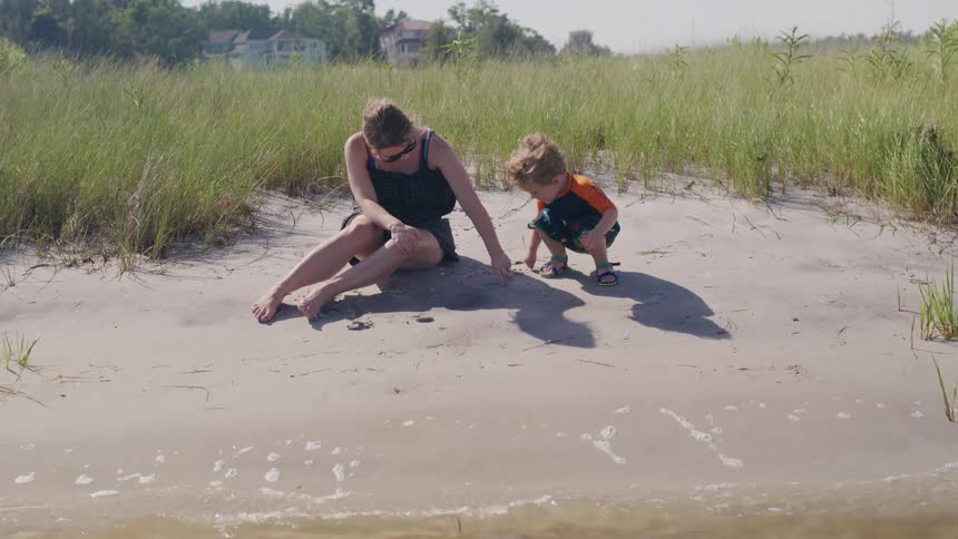 Woman sitting on the sand watches a child playing near the water