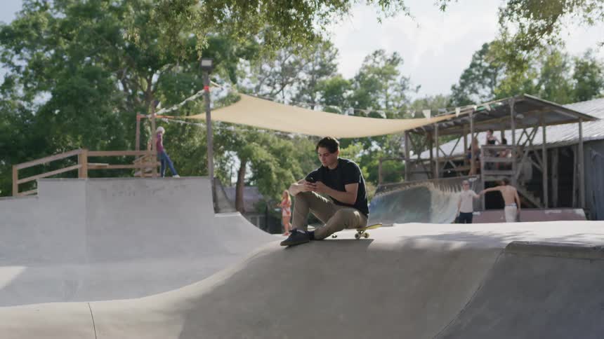 Young man sitting at a skatepark uses his phone with the skateboard under him