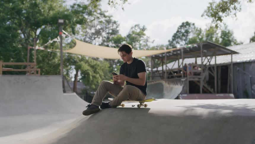 The teenager is using his cell phone while sitting on a skateboard