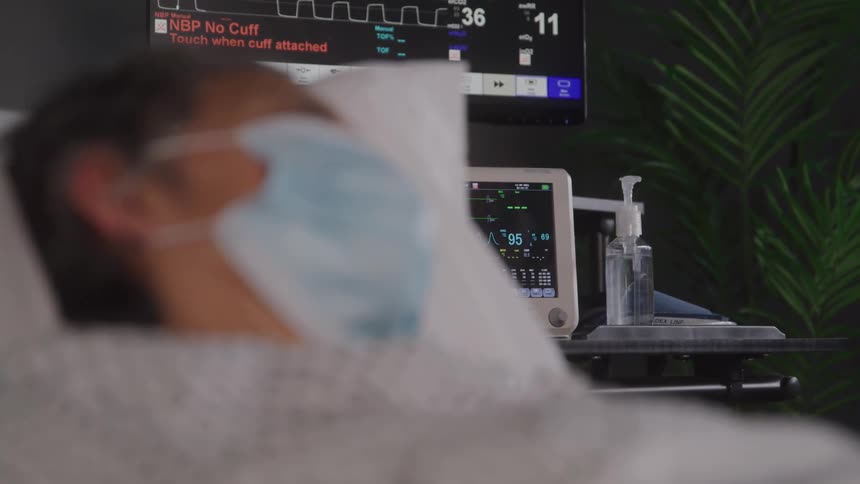A blurry person wearing a face mask lies in a hospital bed with a medical monitor displaying vital signs and a bottle of hand sanitizer on a nearby table