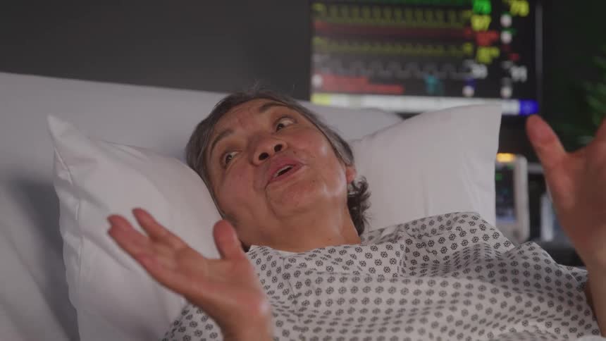 An elderly woman lies on a hospital bed talking with excitement