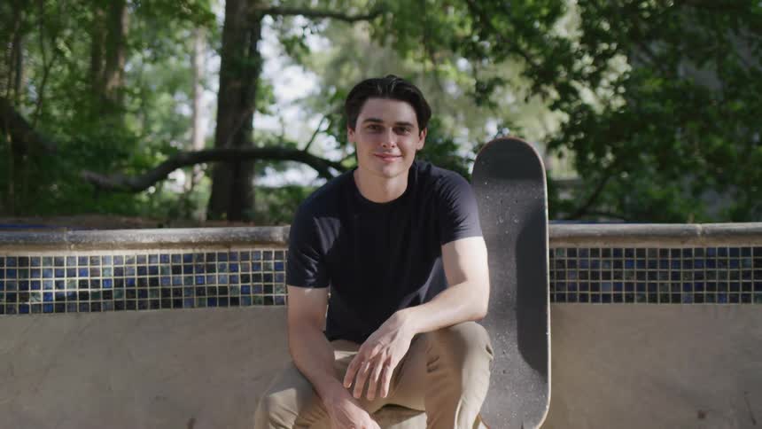 Young man in a black tshirt sits next to a skateboard outdoors