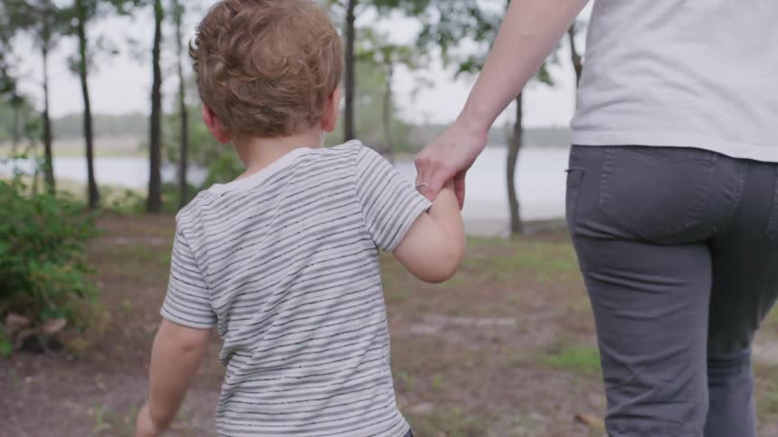 Child in a striped shirt walks hand in hand with an adult in a wooded area