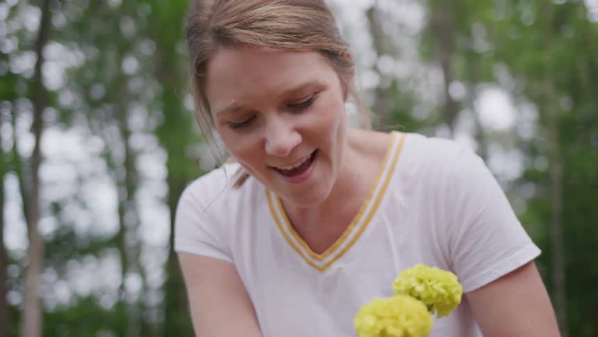 A woman with her hair tied back in a ponytail and a white t shirt with a yellow stripe smiles while planting a small yellow flower she holds in her hands