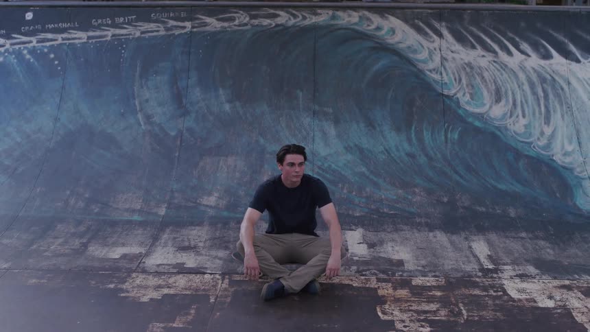 A young man wearing a black tshirt sits on a skateboard in a skateboarding park at midday