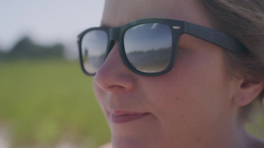 A woman wearing dark sunglasses with a reflection of a landscape in the lenses stands outdoors on a sunny day