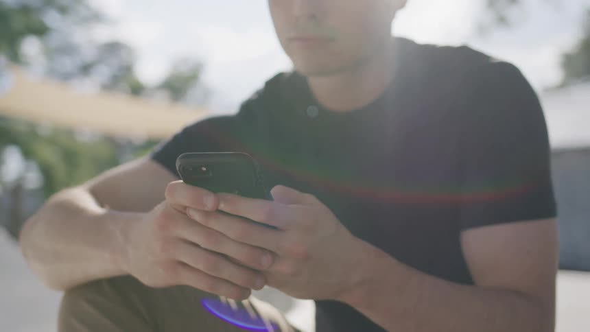 The video shows a closeup of a young man holding a phone and looking down at the screen the sun is shining brightly behind him creating a flare effect on the lens