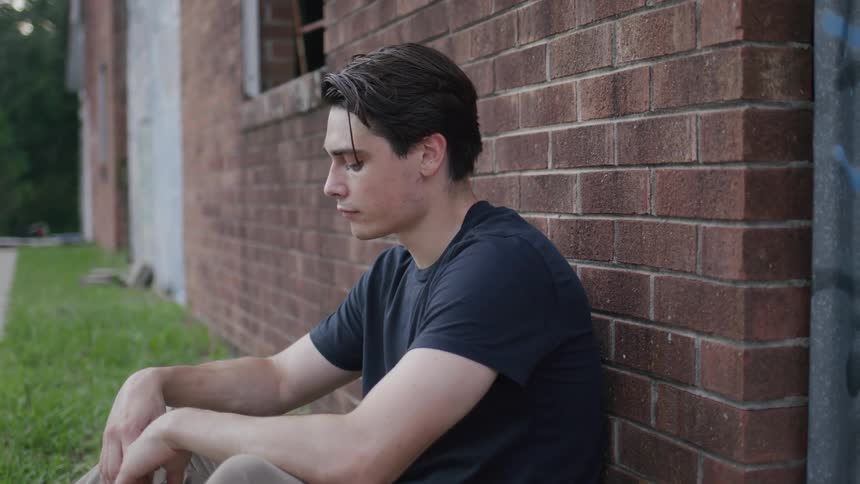 Man in a black tshirt sits on the ground leaning against a brick wall