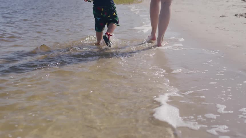 The video shows a closeup of a mother and her child running barefoot in the shallow water along a sandy beach the waves are gently rolling onto the shore and their feet are splashing through the foam