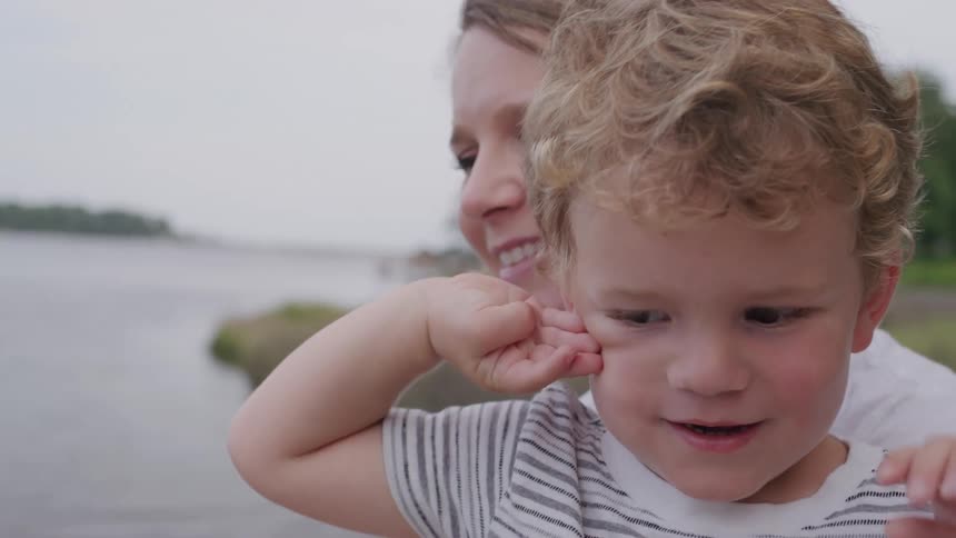 The video shows a child with curly blonde hair and a striped shirt standing outside with an adult the child is looking at something offscreen with their mouth slightly open as if speaking or in awe