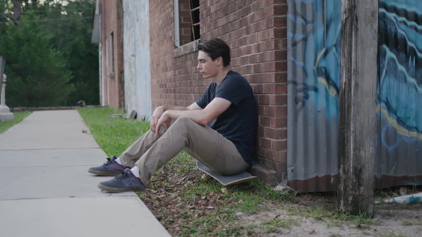 A young male adult sits on his skateboard outdoors beside a building during daytime