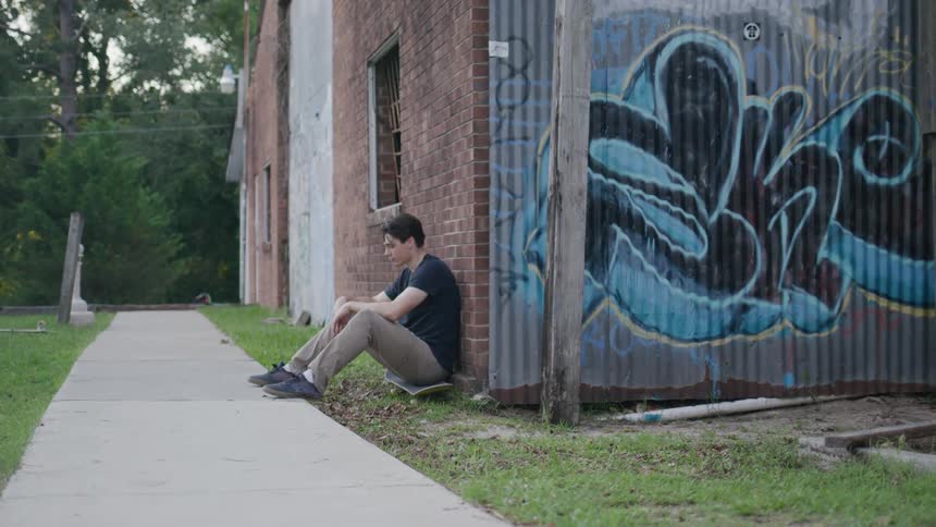 Young man sitting on the ground against a graffitied wall holds a skateboard