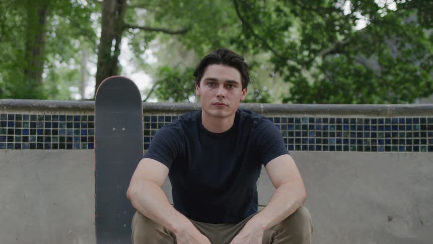 A male skateboarder in a black shirt and khaki pants is standing on a ledge next to an empty skate bowl looking intently into the camera