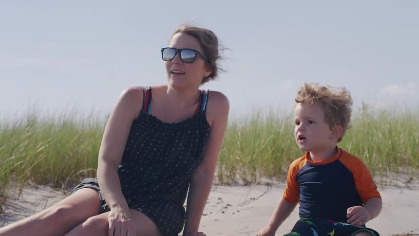 The video shows a mother and her young child sitting on a sandy beach next to tall grass the mother is wearing sunglasses and a black swimsuit and the child is looking at her with a surprised expression as she points offscreen and talks
