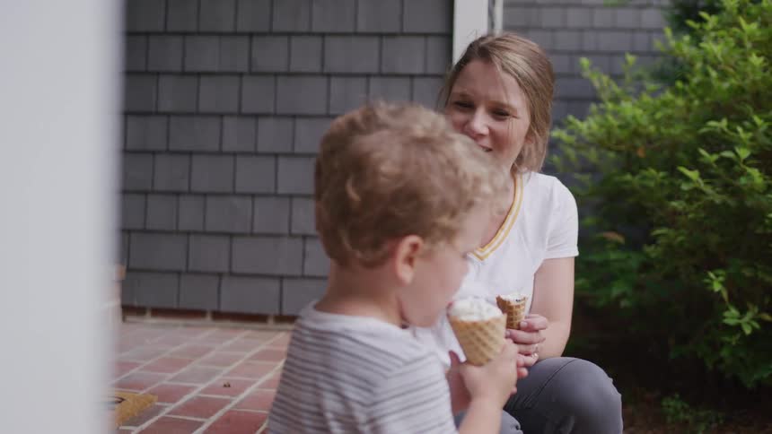 A woman and her toddler sit on their front porch both holding ice cream cones and having fun at midday