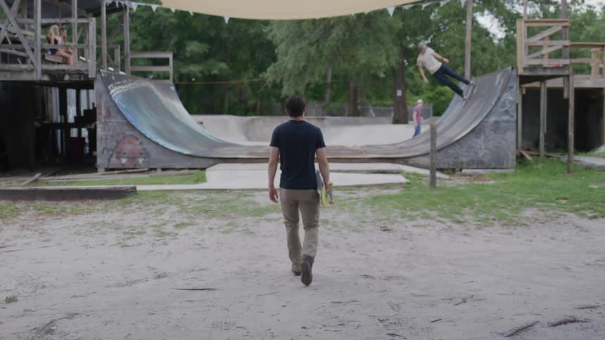 Young man walks with a skateboard in hand toward a wooden ramp