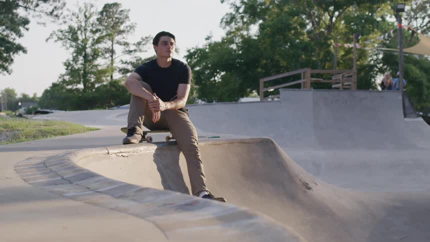 A man in a black shirt and brown pants sits on the edge of a concrete skate bowl resting over his skateboard