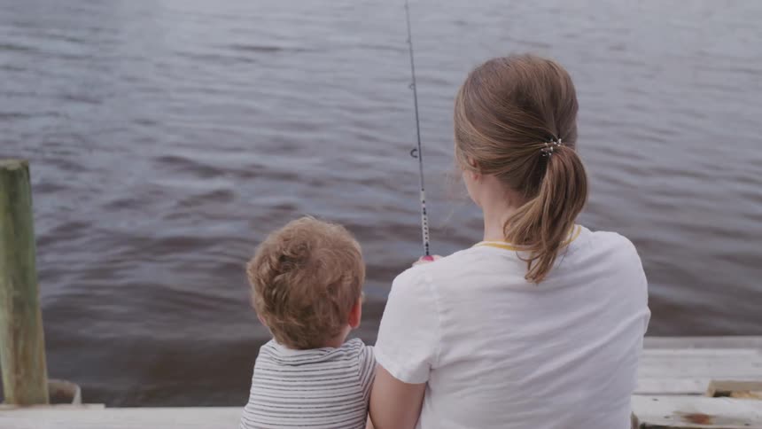 A woman and a young child are sitting on a wooden dock facing away from the camera and looking out over a body of water while holding a fishing rod