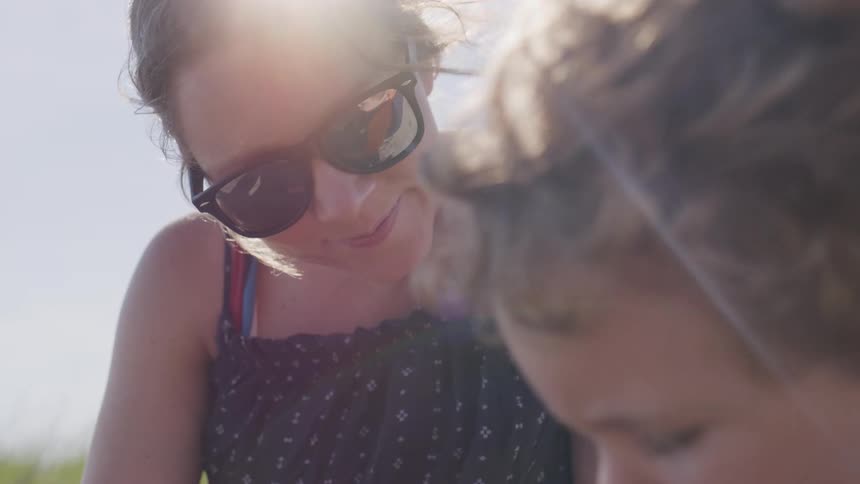 Woman wearing sunglasses looks at a child under bright sunlight in an outdoor setting