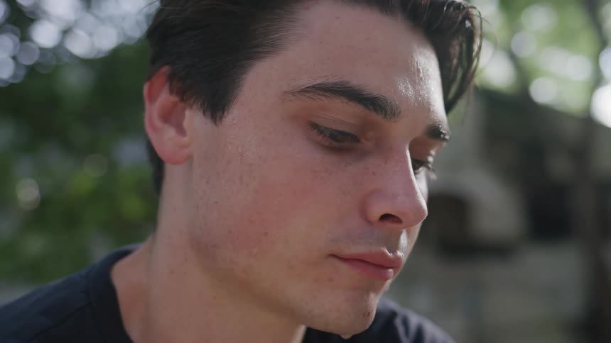 A young man with dark hair and a serious expression is looking down and to the side the shot is a closeup on his face with blurred greenery visible in the background