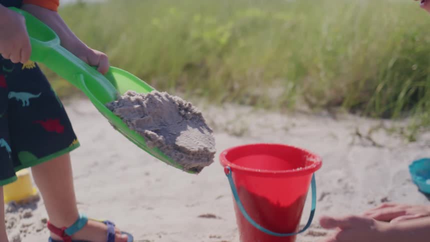 A child digs in the sand with a green shovel beside a womans hand and a red bucket