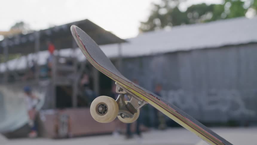 A skateboard is held up in focus at a skate park with ramps and skaters blurred in the background