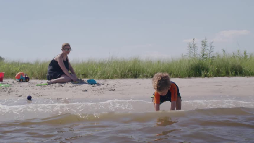 A woman sits on the sand while a small boy wearing a long sleeved swim shirt and shorts plays in the water nearby