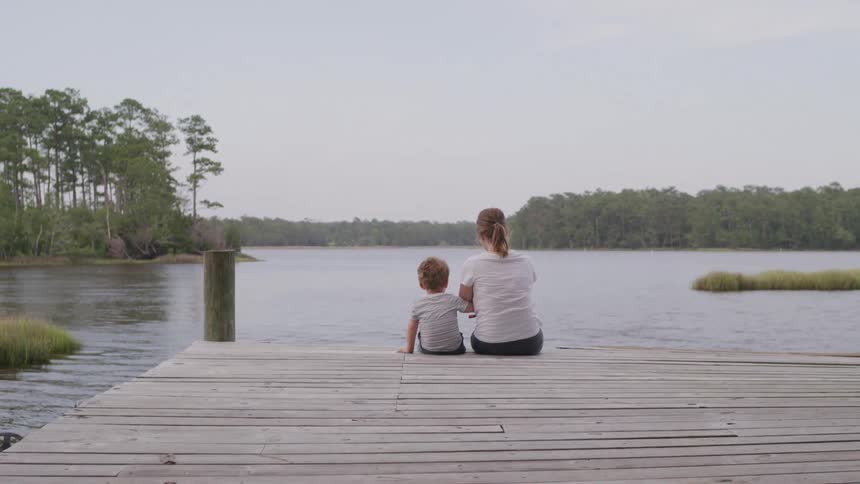A woman sits at a dock with her son beside her fishing at midday