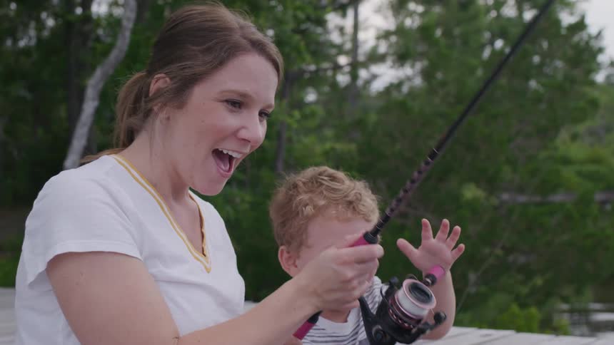 A joyous closeup shot of a woman and a young child on a dock smiling and laughing as they hold a fishing rod together