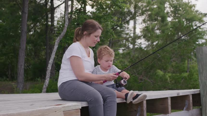 Mother teaches her son how to fish while sitting on the dock near a grove of trees