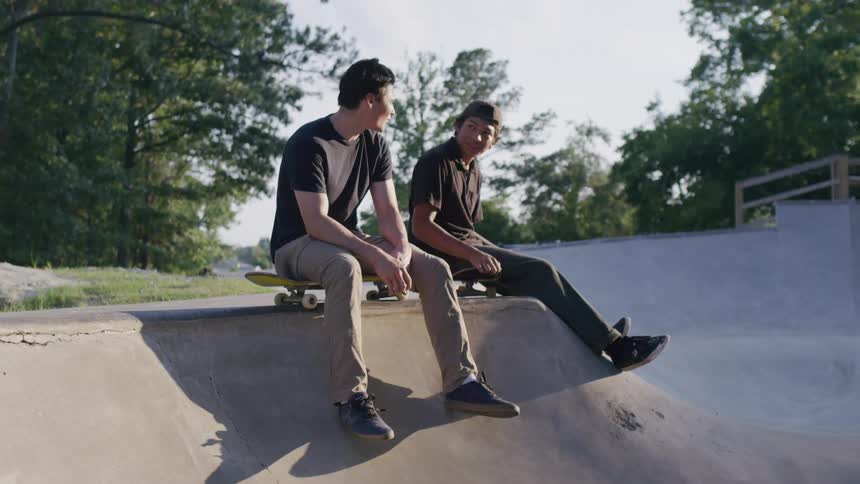 Two young men talk while sitting with skateboards at a concrete skatepark