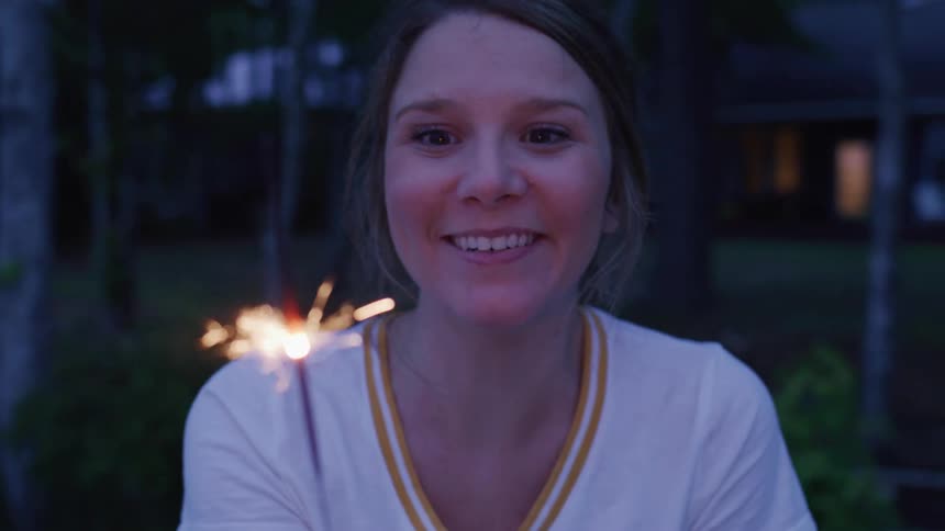 A woman with brown hair smiles while holding a lit sparkler in front of her face with a blurry nighttime background