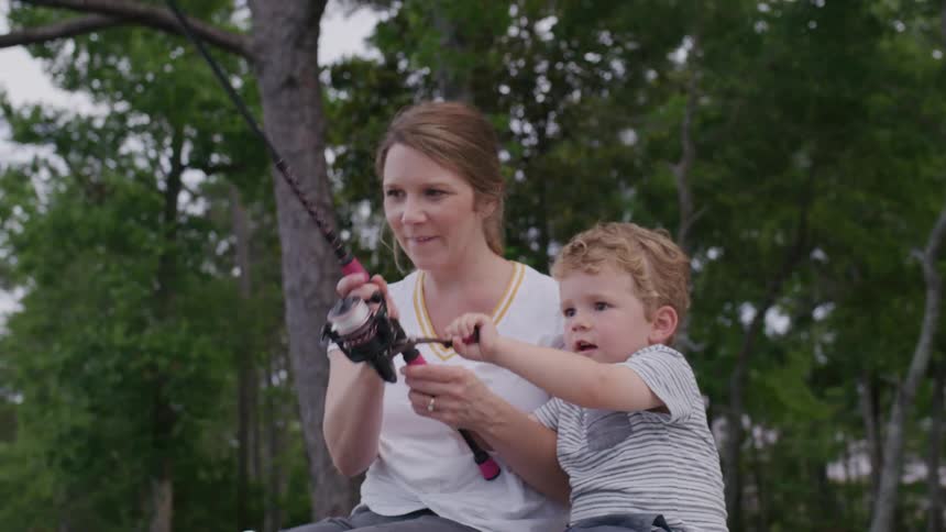 A young boy and his mother sit on a bench outdoors as they fish together with the boy holding the fishing pole and his mother guiding his hands