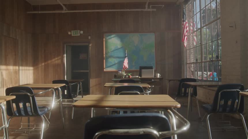 An empty classroom with a series of desks and chairs features a map on the board and a desk with an american flag at midday