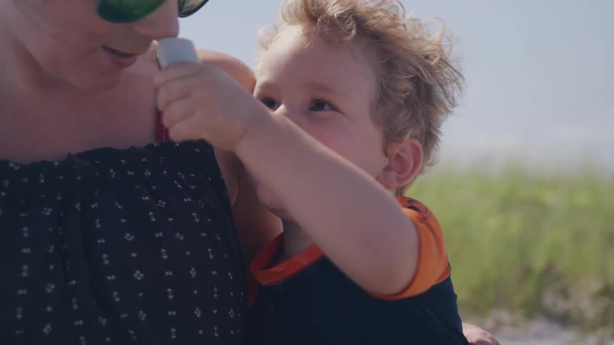 The video shows a mother applying sunscreen to a young boys nose who is looking up at her with a sweet and slightly curious expression they are outdoors on what appears to be a beach with sand and a hint of the sea in the background