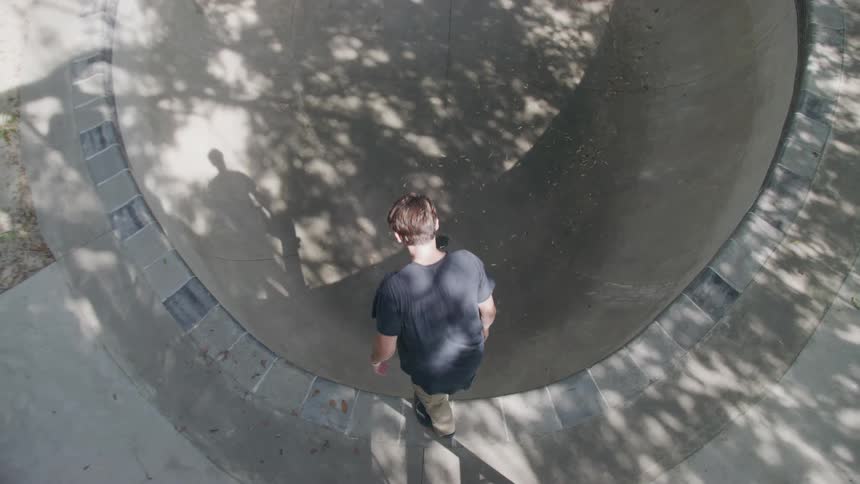 A teenager wearing a dark t shirt and light pants slides down a concrete skate bowl on a sunny day with dappled light on the ground