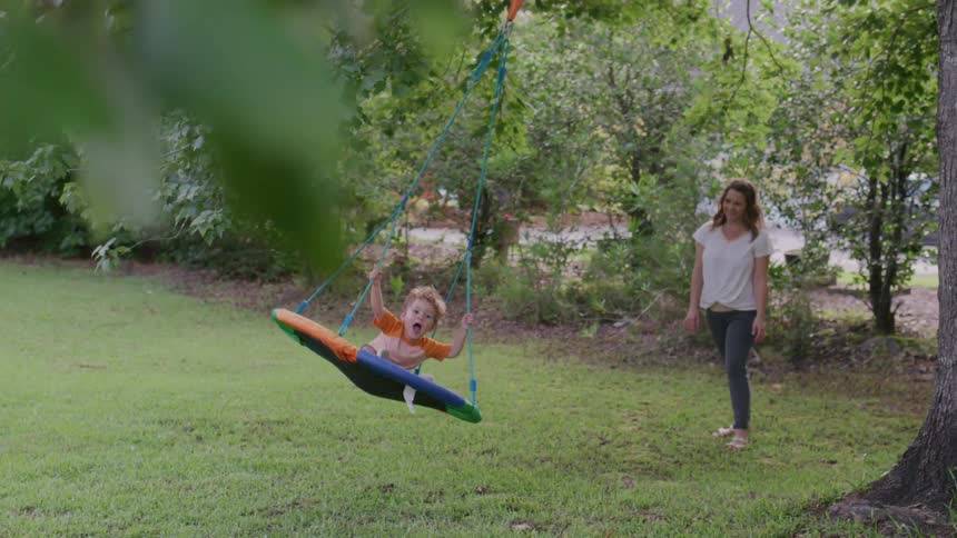 A mother gently pushes her young child on a round swing in a grassy backyard with trees and foliage in the background