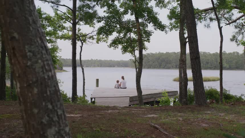 A mother and child sit together at the end of a dock looking out over a calm lake