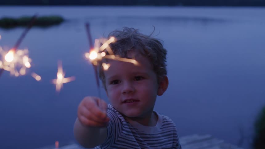 A young boy holds a lit sparkler near a lake at dusk
