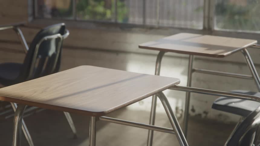 Empty student desks and chairs are arranged in a classroom with sunlight streaming through the windows and casting shadows on the floor