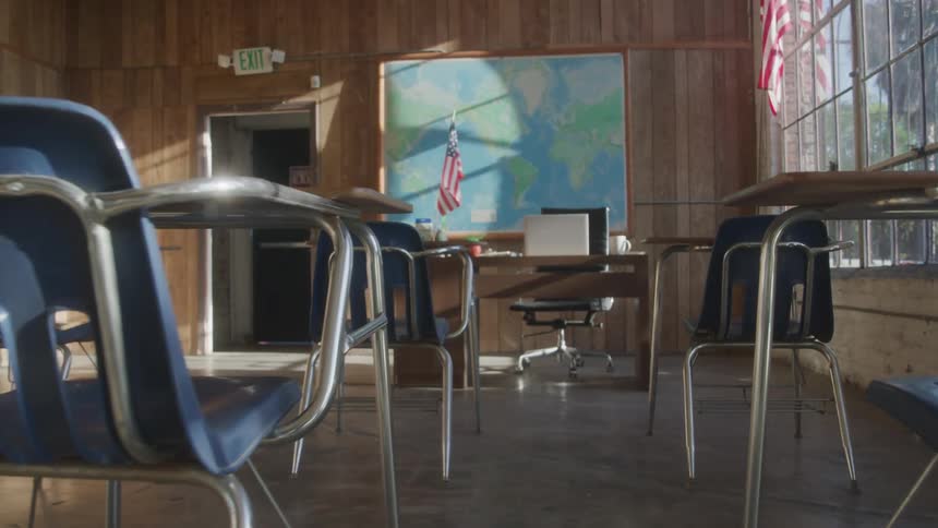 A sunny classroom is empty except for the student desks and chairs a teachers desk with a laptop and a large map on a wood paneled wall